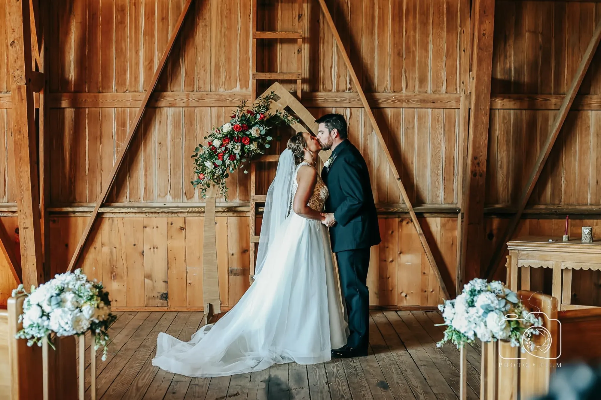 Bride and groom sharing a first kiss beneath a wooden arch with floral arrangements